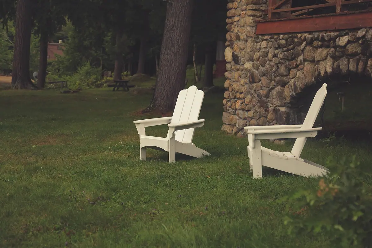 Two white Adirondack chairs on a green lawn beside a stone wall and tall pines