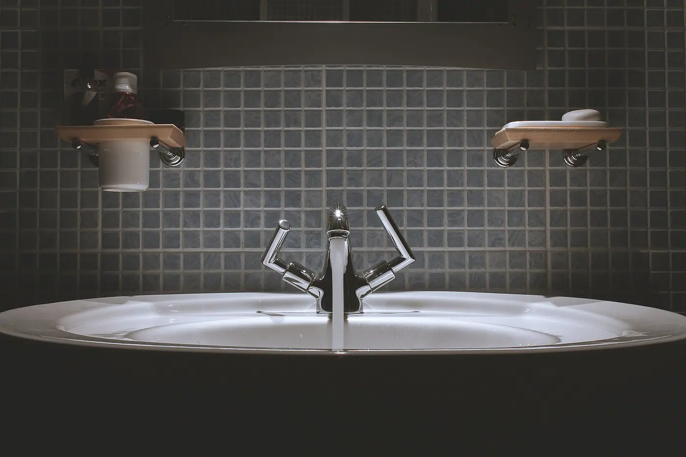 Chrome faucet running water into a white basin against dark mosaic tile