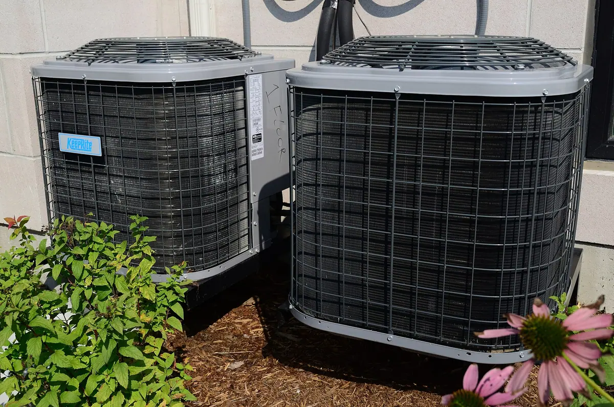 Two residential air conditioner condenser units beside a house with landscaping