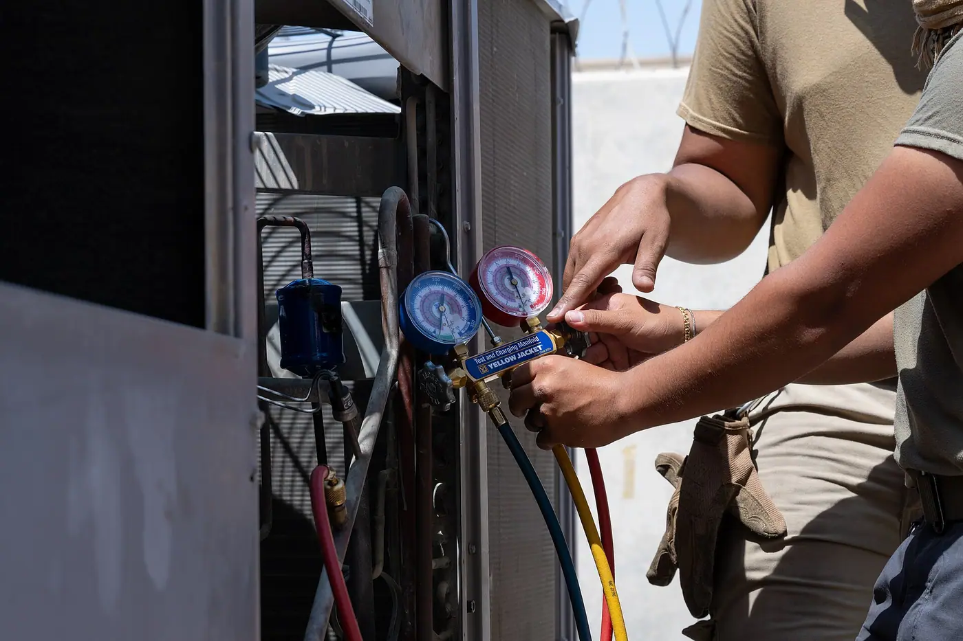 Technician's hands connecting a refrigerant manifold gauge set with red, blue and yellow hoses to an air conditioning unit