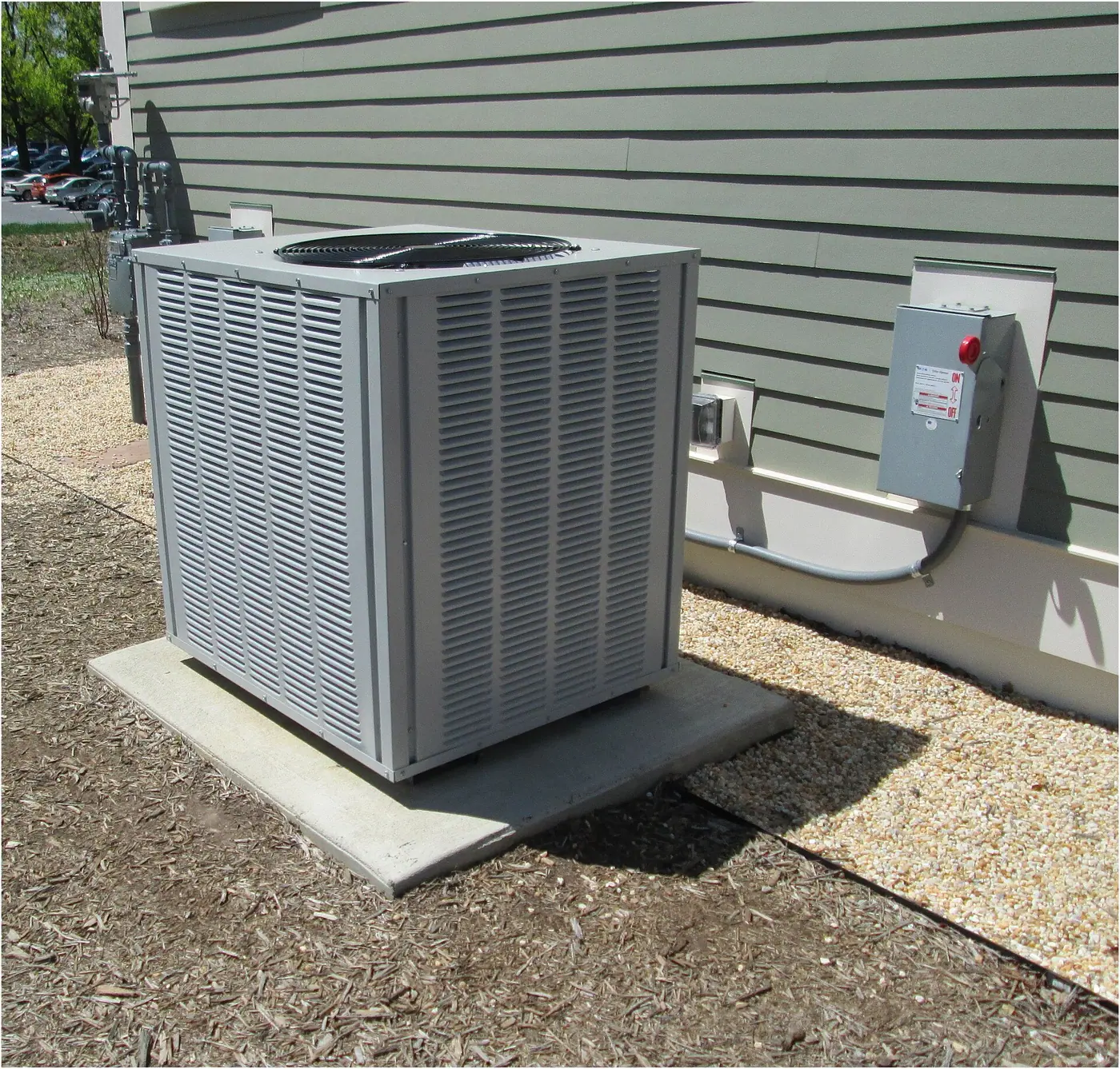 Residential central air conditioner condenser on a concrete pad beside green lap siding with a disconnect box
