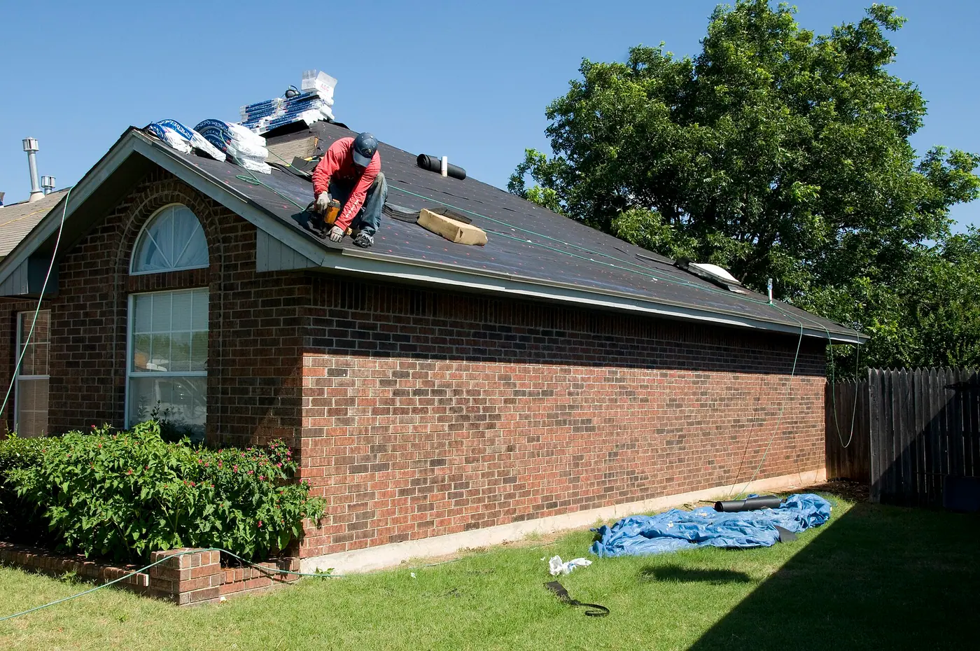Roofer nailing new shingles on the roof of a brick ranch house under a clear blue sky